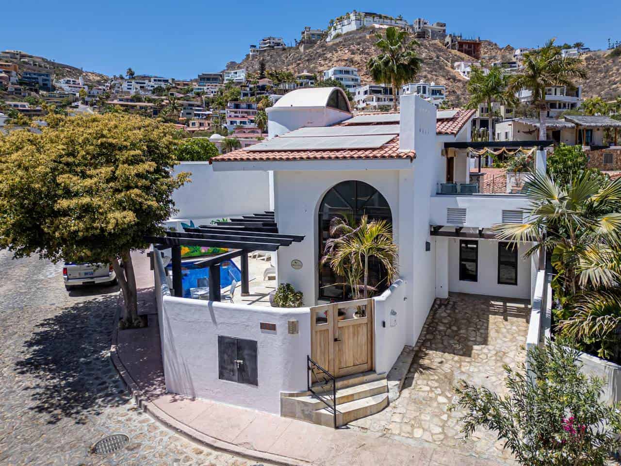 Modern White Pedregal Home With A Red-Tile Roof, Surrounded By Lush Palm Trees And Rocky Hillside Views, Showcasing Contemporary Design And Scenic Coastal Living In Pedregal.