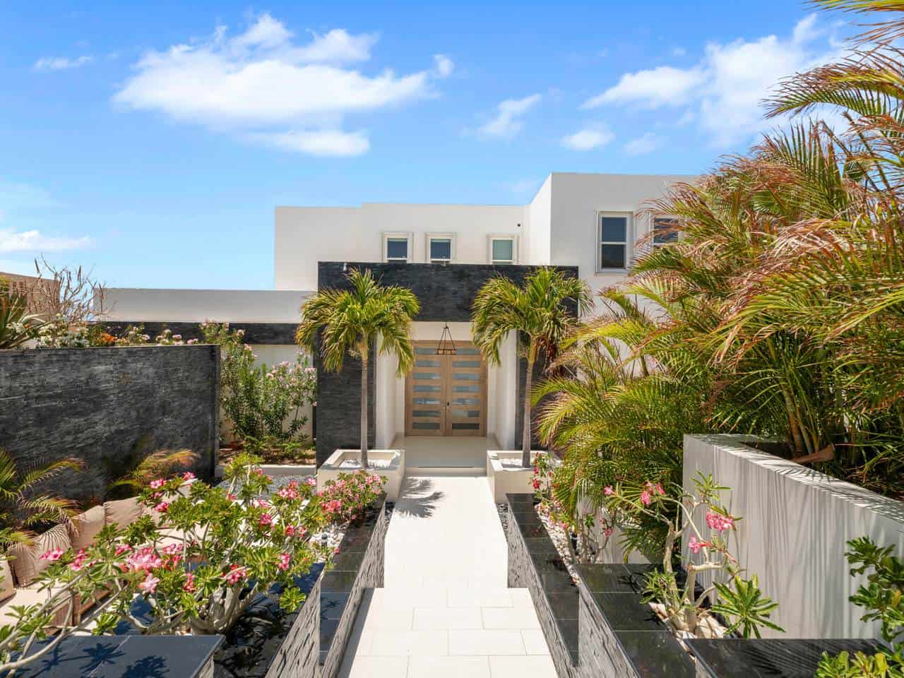 Luxurious Pedregal Modern Home Entrance With Lush Tropical Landscaping And Vibrant Flowers Under A Bright Blue Sky.