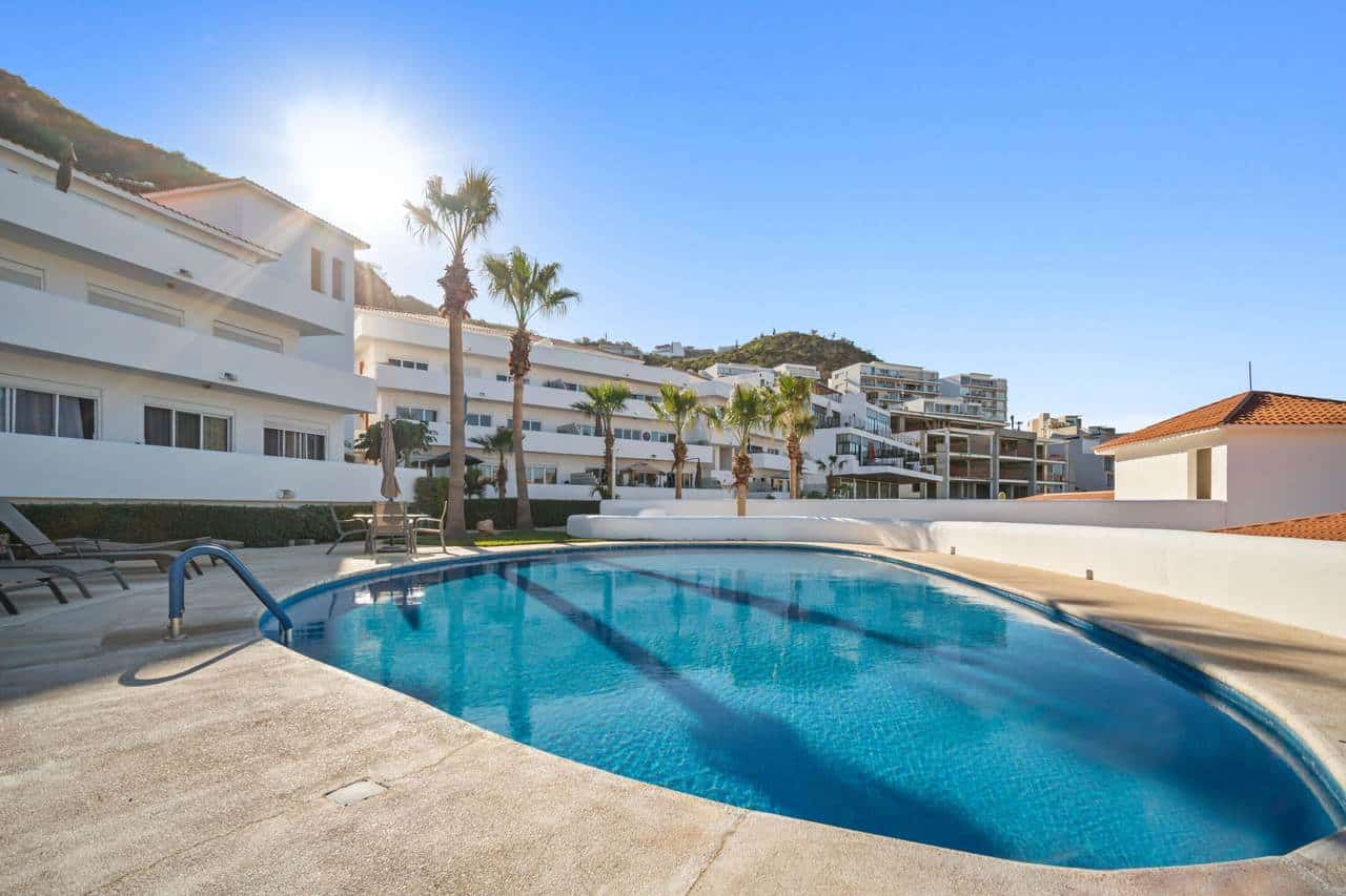 Luxury Pedregal Condo With Swimming Pool And Ocean Views, Modern White Buildings, Tall Palm Trees, And Hillside Background, In A Sunny Coastal Setting.